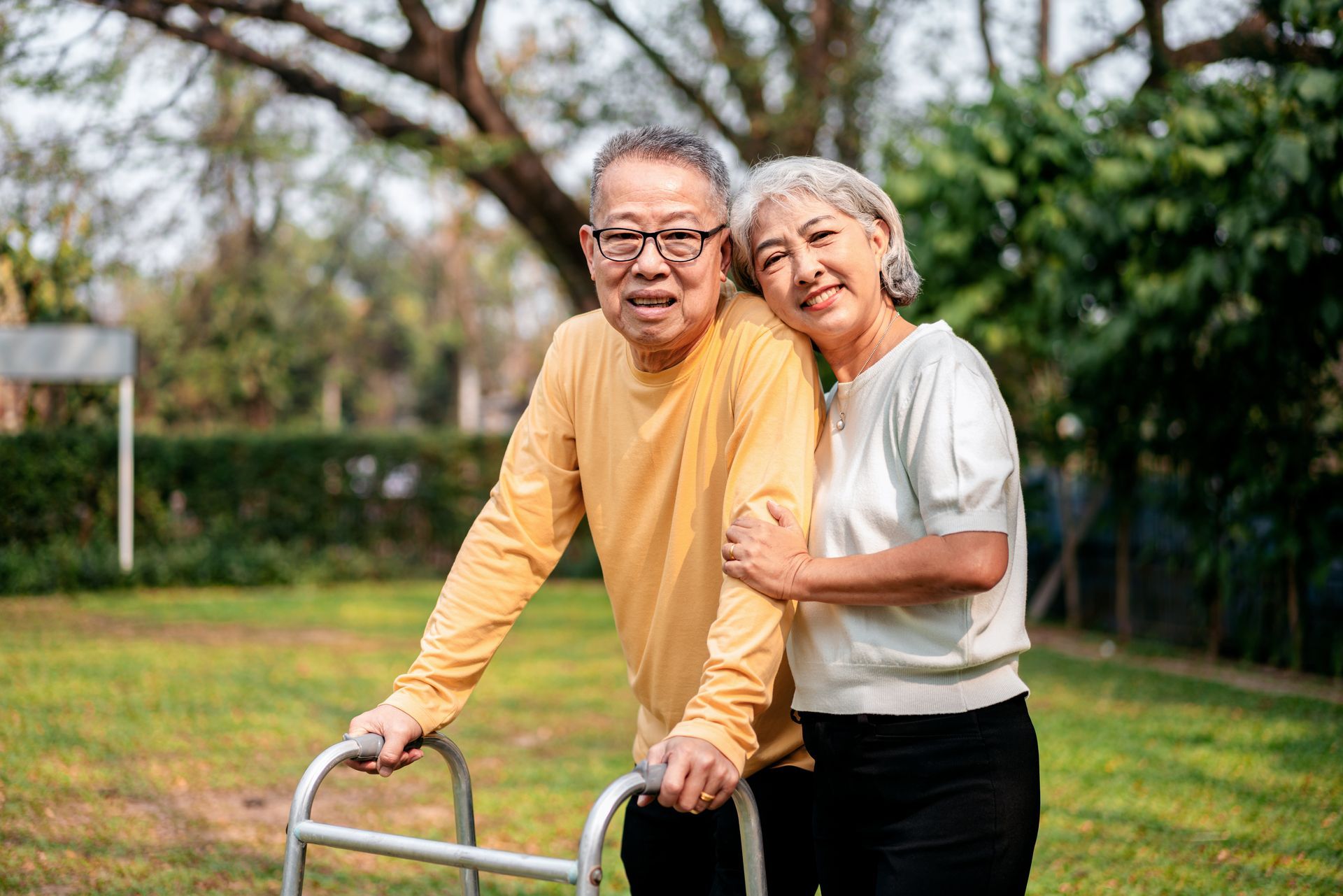 Elderly couple outdoors, one using a walker, other has arm around their shoulder, looking at the camera.