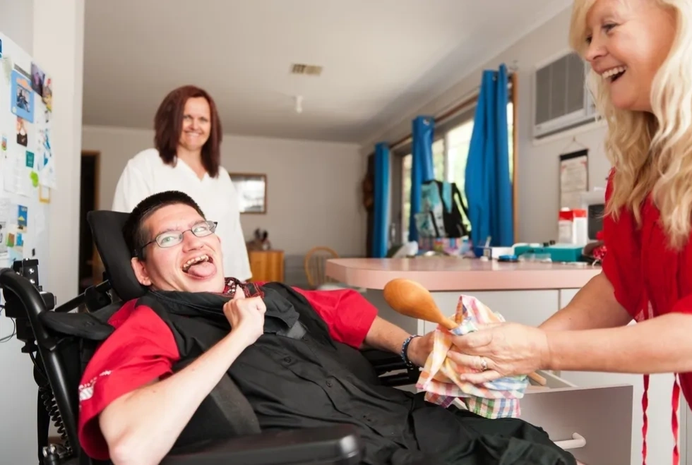 Man in wheelchair smiles, interacting with a woman. Another woman watches in a home kitchen setting.