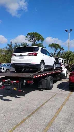 White SUV on a flatbed tow truck in a parking lot on a sunny day.