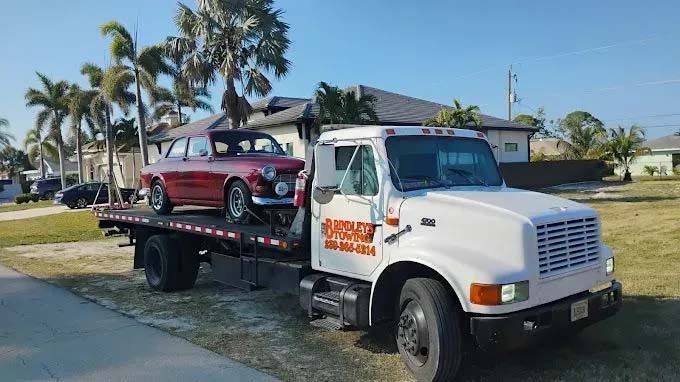 Red classic car on a flatbed tow truck, white truck parked on grass. Suburban setting.