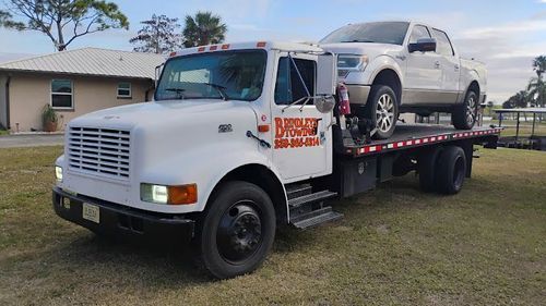 White tow truck with a white pickup truck on its flatbed, parked in front of a house.
