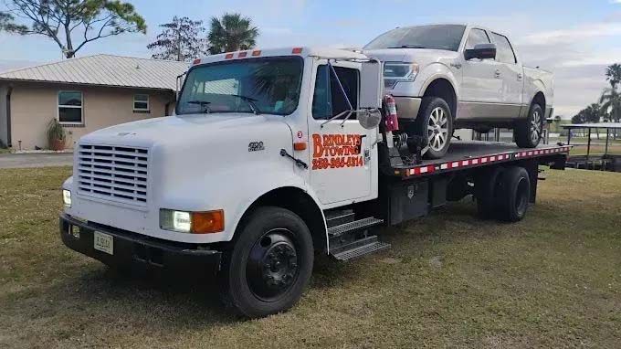 White tow truck carrying a white pickup truck on a flatbed. Grass and a house in the background.