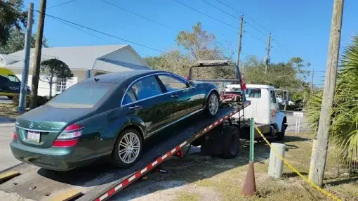 Green Mercedes sedan being loaded onto a flatbed tow truck on a sunny day.