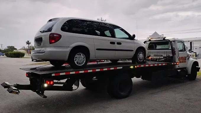 A silver minivan being towed on a flatbed tow truck, outdoors, under a cloudy sky. A silver minivan being towed on a flatbed tow truck, outdoors, under a cloudy sky.