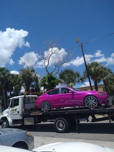 A pink car with chrome rims on a tow truck, under a bright blue sky with palm trees.