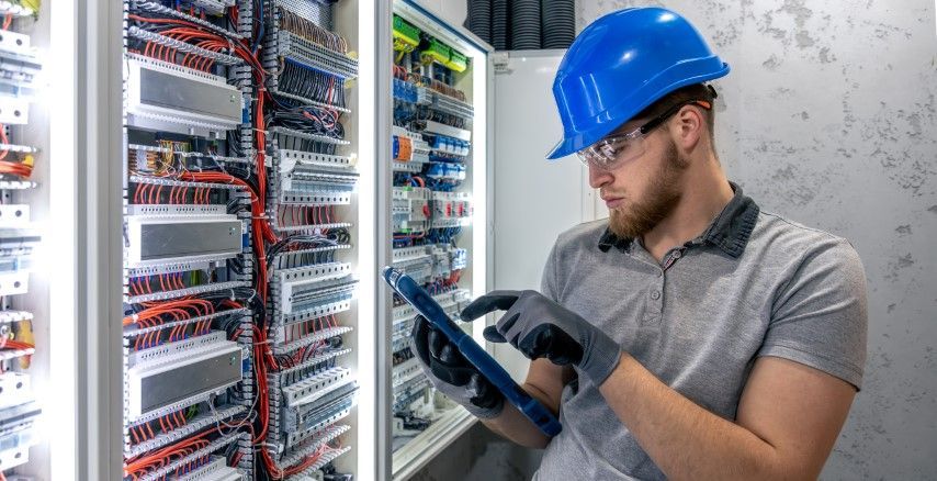 Electrician in blue hard hat and gloves inspecting electrical panel with tablet.
