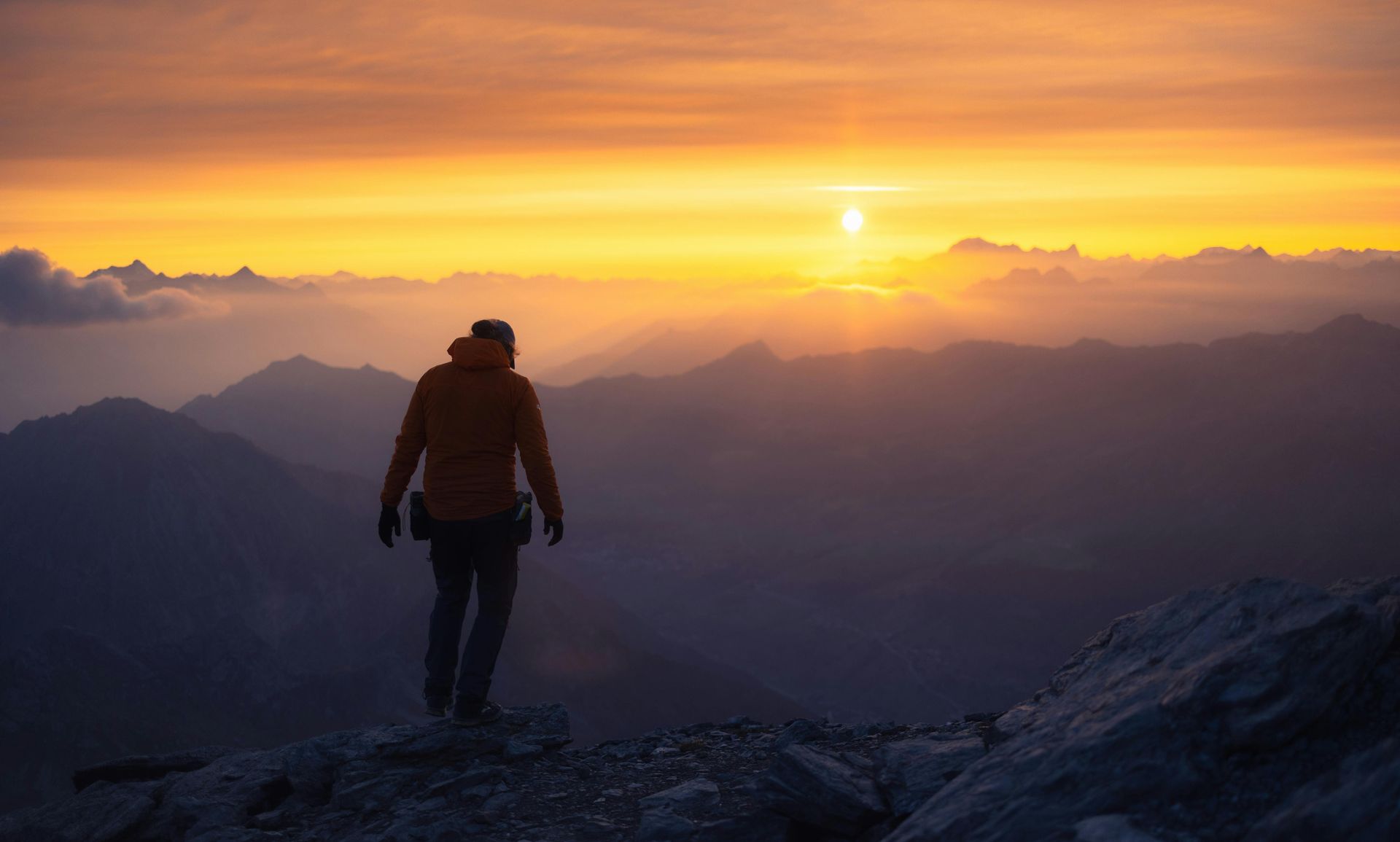 A person is standing on top of a mountain looking at the sunset.