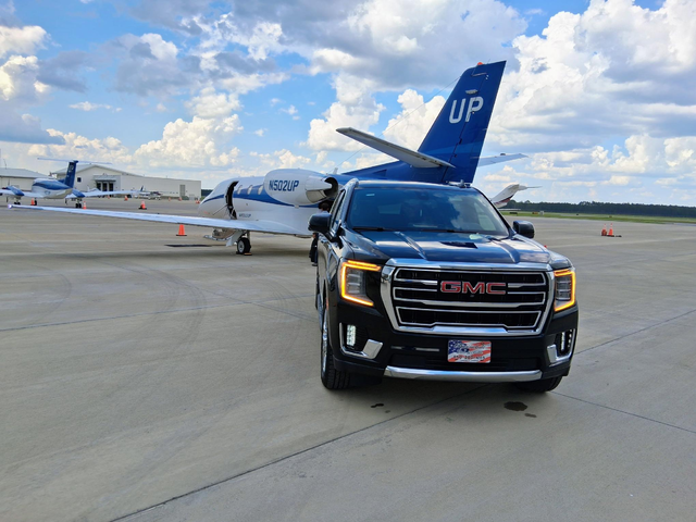Black GMC Yukon Denali parked on a tarmac, private jet in background, blue sky.