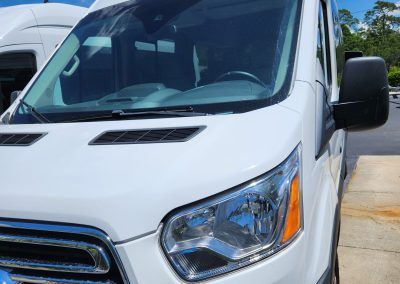 White van's front, driver inside. Headlight, side mirror, and windshield wipers visible. Parked outside.