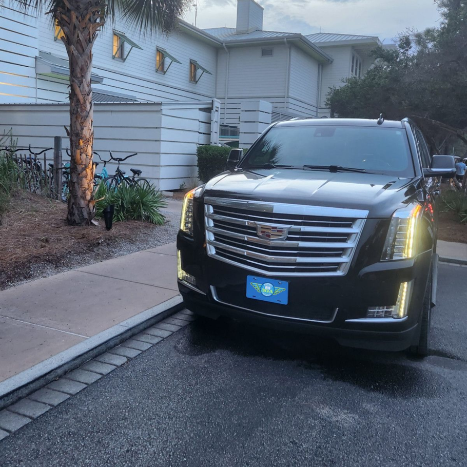 Black Cadillac SUV parked in front of a white building with a palm tree, bikes, and overcast sky.