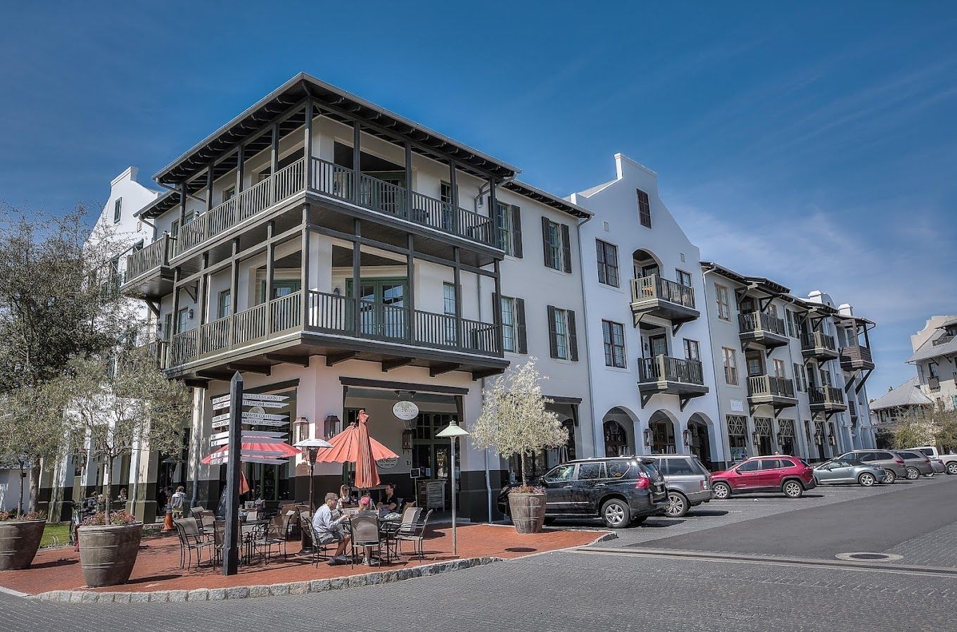 Multi-story white building with balconies, street-side restaurant with patrons, parked cars, clear sky.