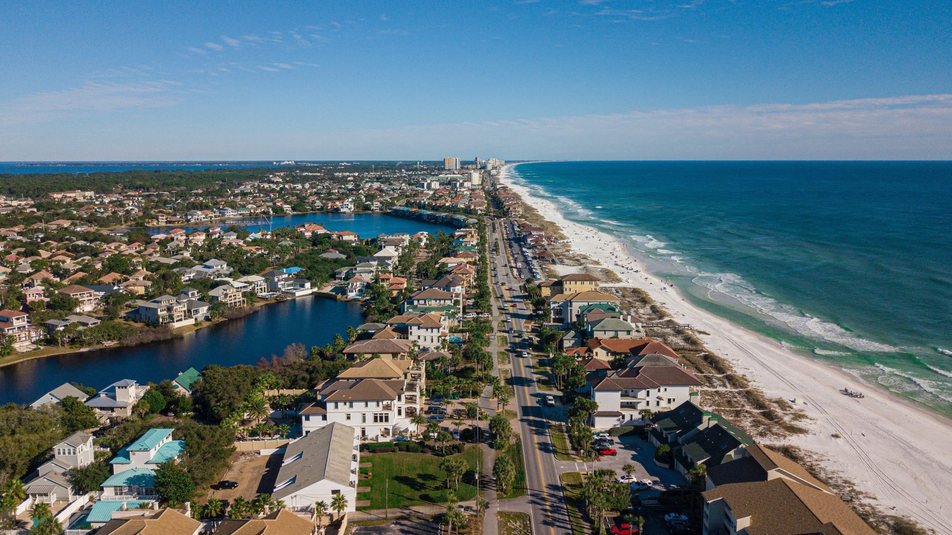 Aerial view of a coastal town with a sandy beach, turquoise water, and houses lining the road.