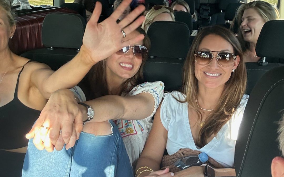 Women in sunglasses smile and wave from inside a vehicle.