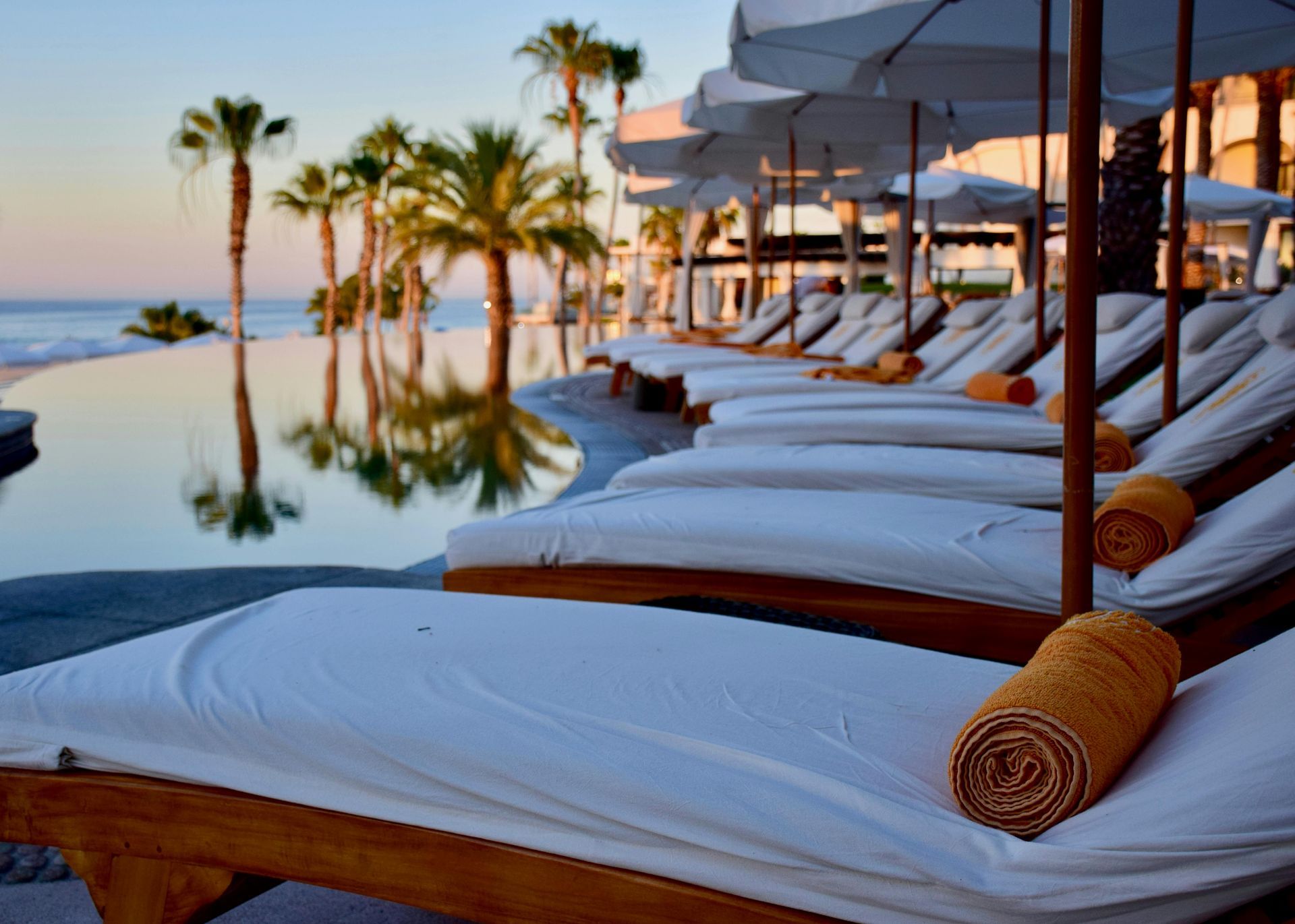 Lounge chairs with rolled towels line a pool overlooking the ocean, palm trees in the background at sunset.