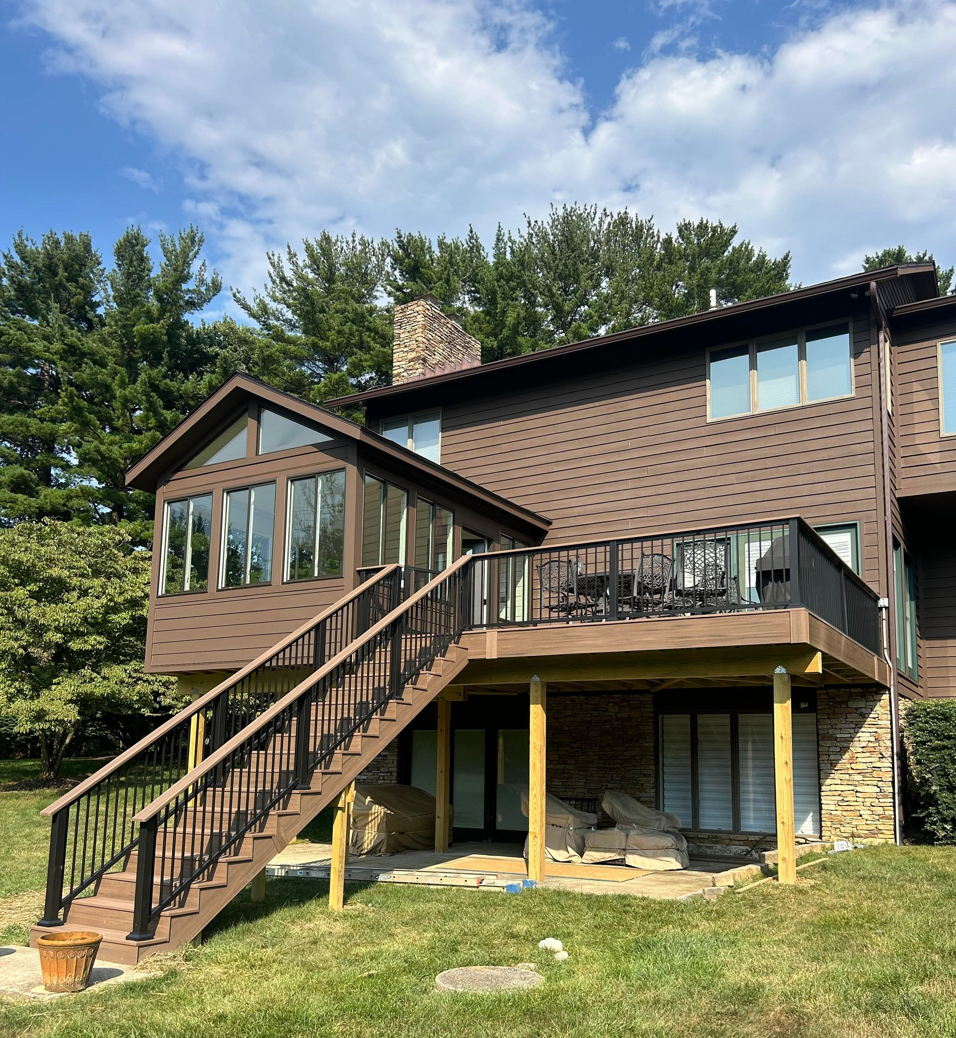 beam wraps Brown house with deck, stairs, and sunroom, set in a grassy backyard. Sunny day.