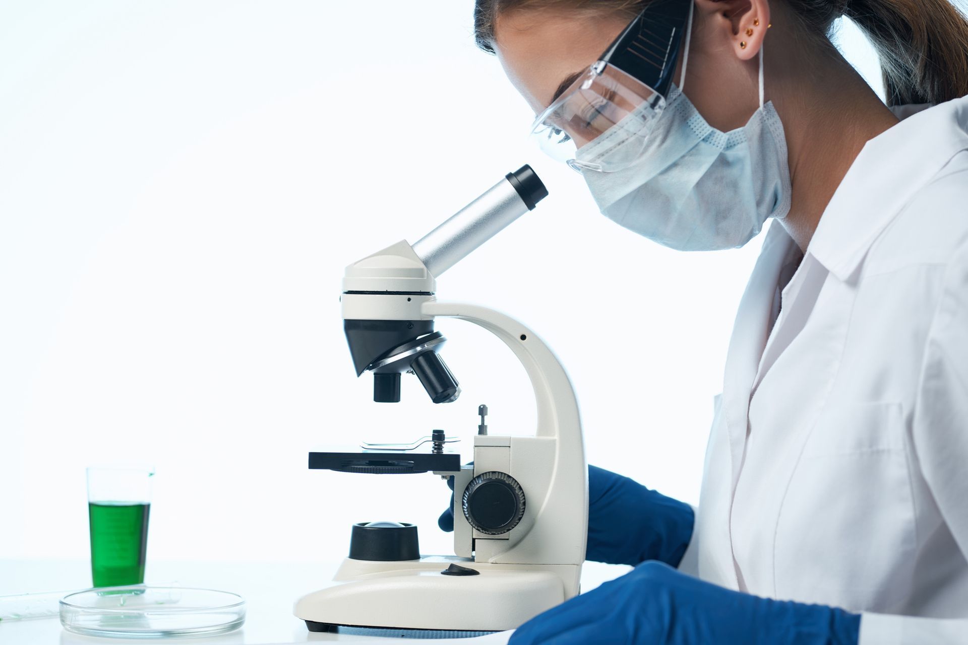 Uno scienziato in camice, occhiali protettivi e maschera esamina un campione al microscopio.