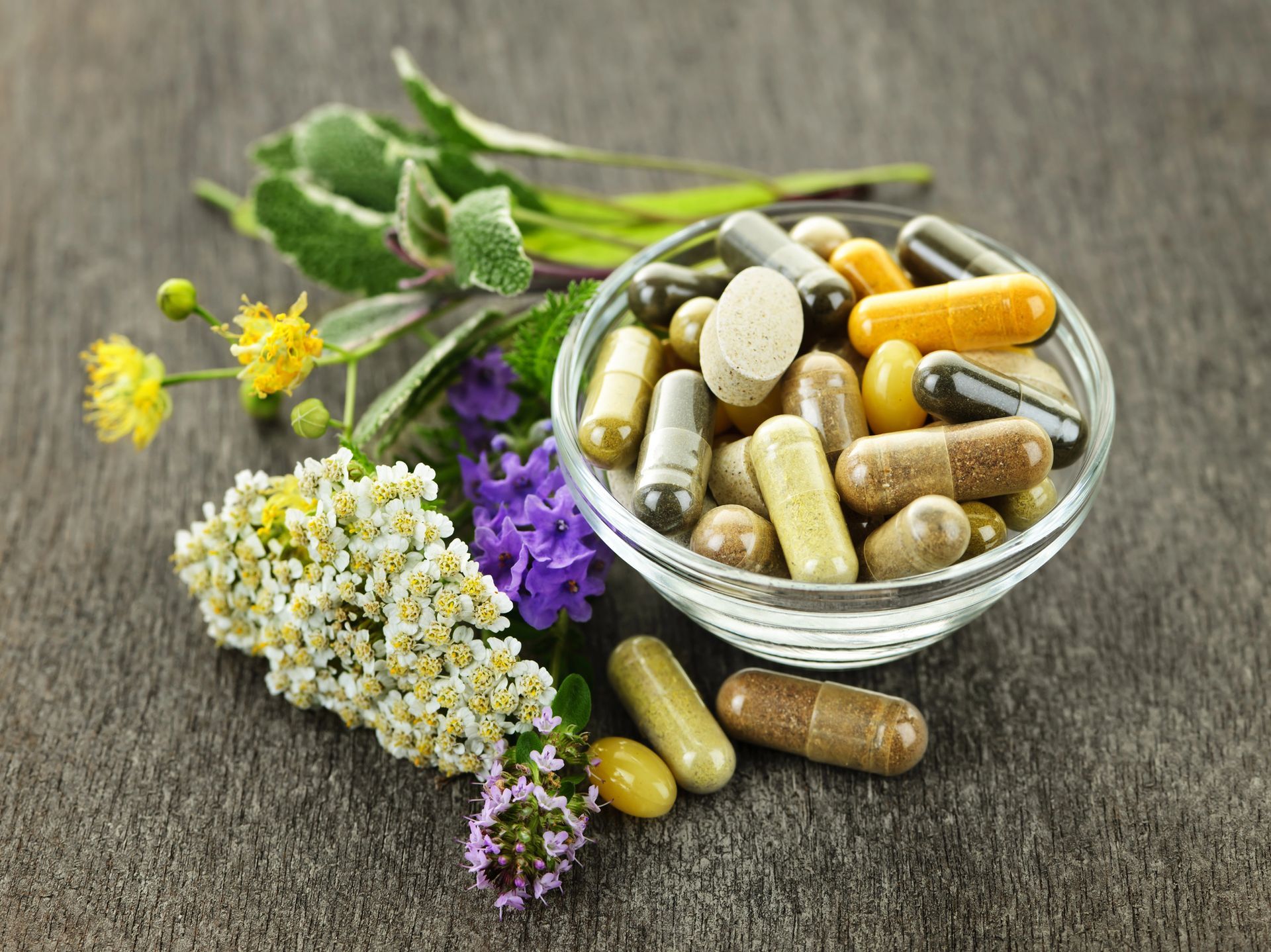 Ciotola di pillole e capsule con erbe e fiori su una superficie di legno.