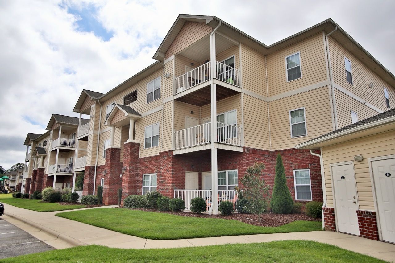 A three-story apartment complex with beige siding, brick accents, and balconies on a grassy lot under a cloudy sky.
