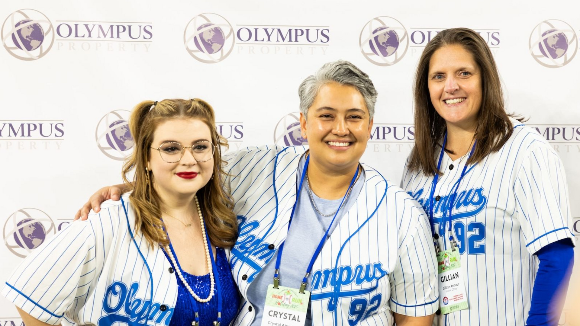 Three smiling people in blue-and-white pinstriped Olympus jerseys standing in front of an Olympus branded backdrop.