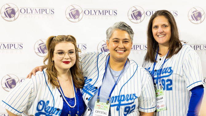 Three smiling people in blue-and-white pinstriped Olympus jerseys standing in front of an Olympus branded backdrop.