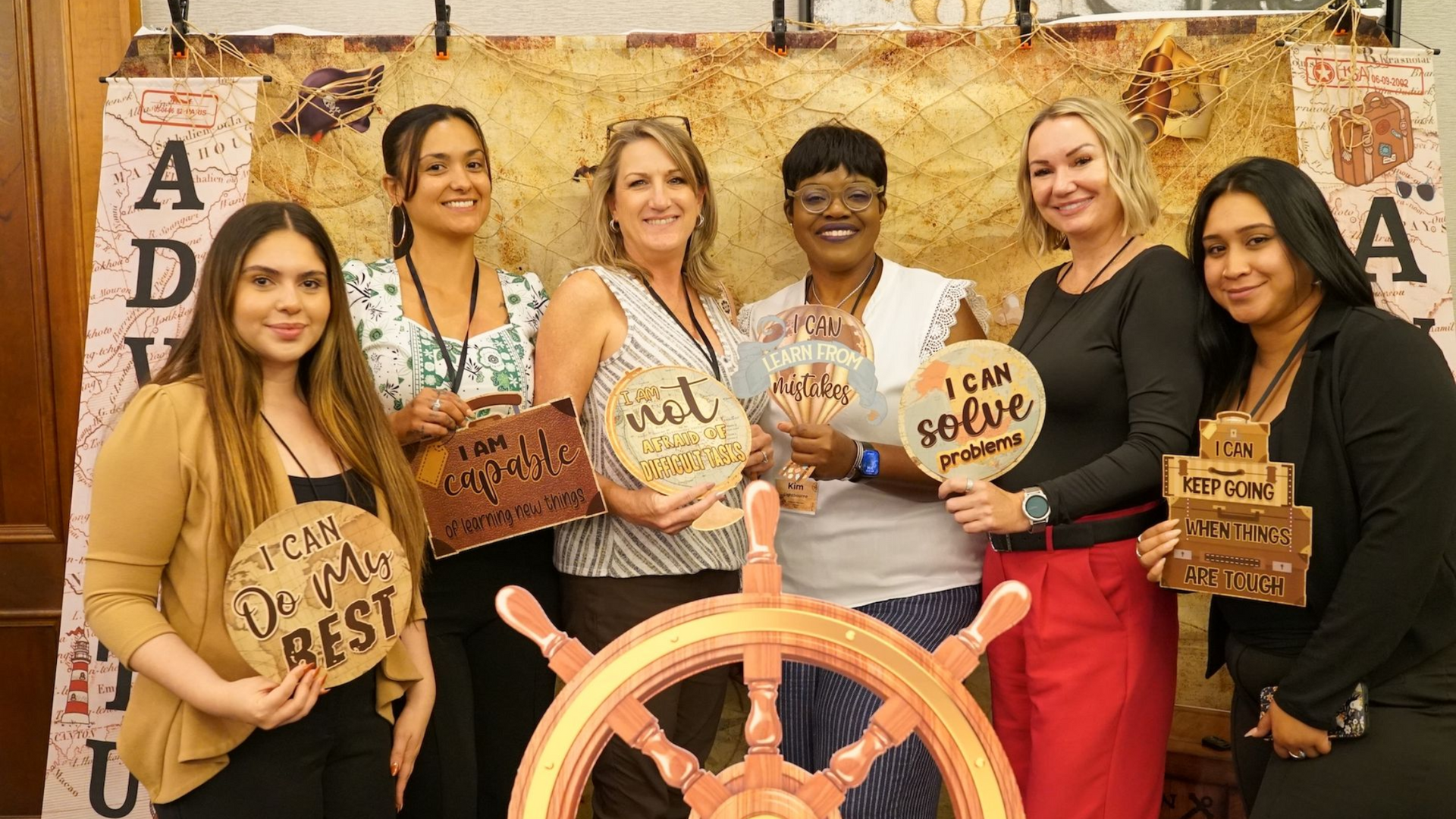 Six people hold signs with positive affirmations in front of a rustic, map-themed background and a ship's wheel.