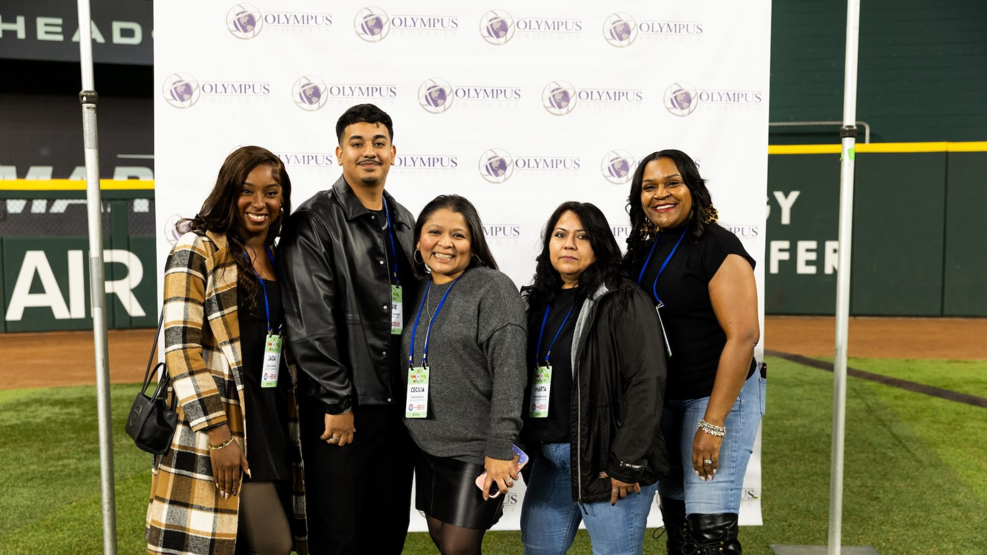 Five people pose for a photo in front of a branded event backdrop on a baseball field.