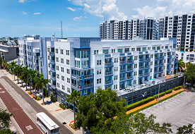 An aerial view of a modern multi-story apartment building with a white and blue exterior, a street, and a parking lot.
