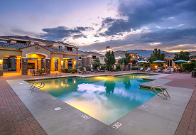 A pool at a desert apartment complex at sunset, with a multi-story building, patio furniture, and lit windows.