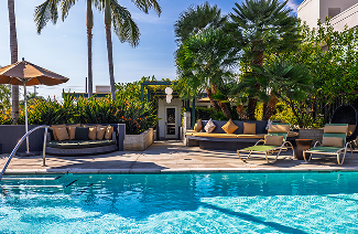 A sunny pool deck with lounge seating, palm trees, and an umbrella by a blue swimming pool.