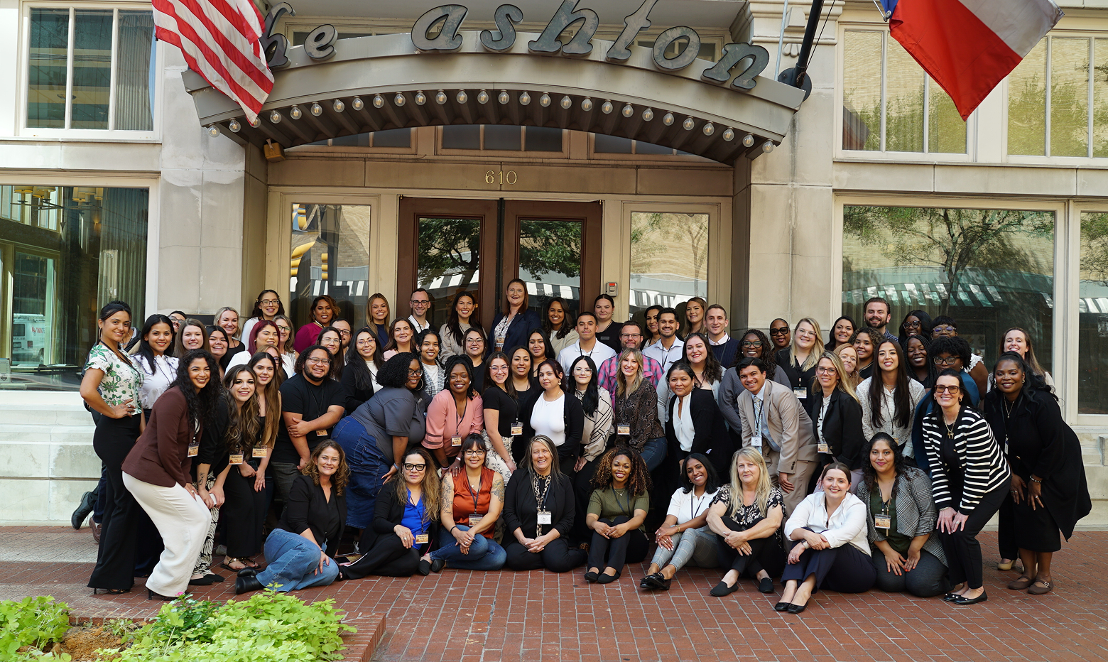 A large group posing for a photo in front of The Ashton hotel entrance, with an American flag and a Texas flag overhead.