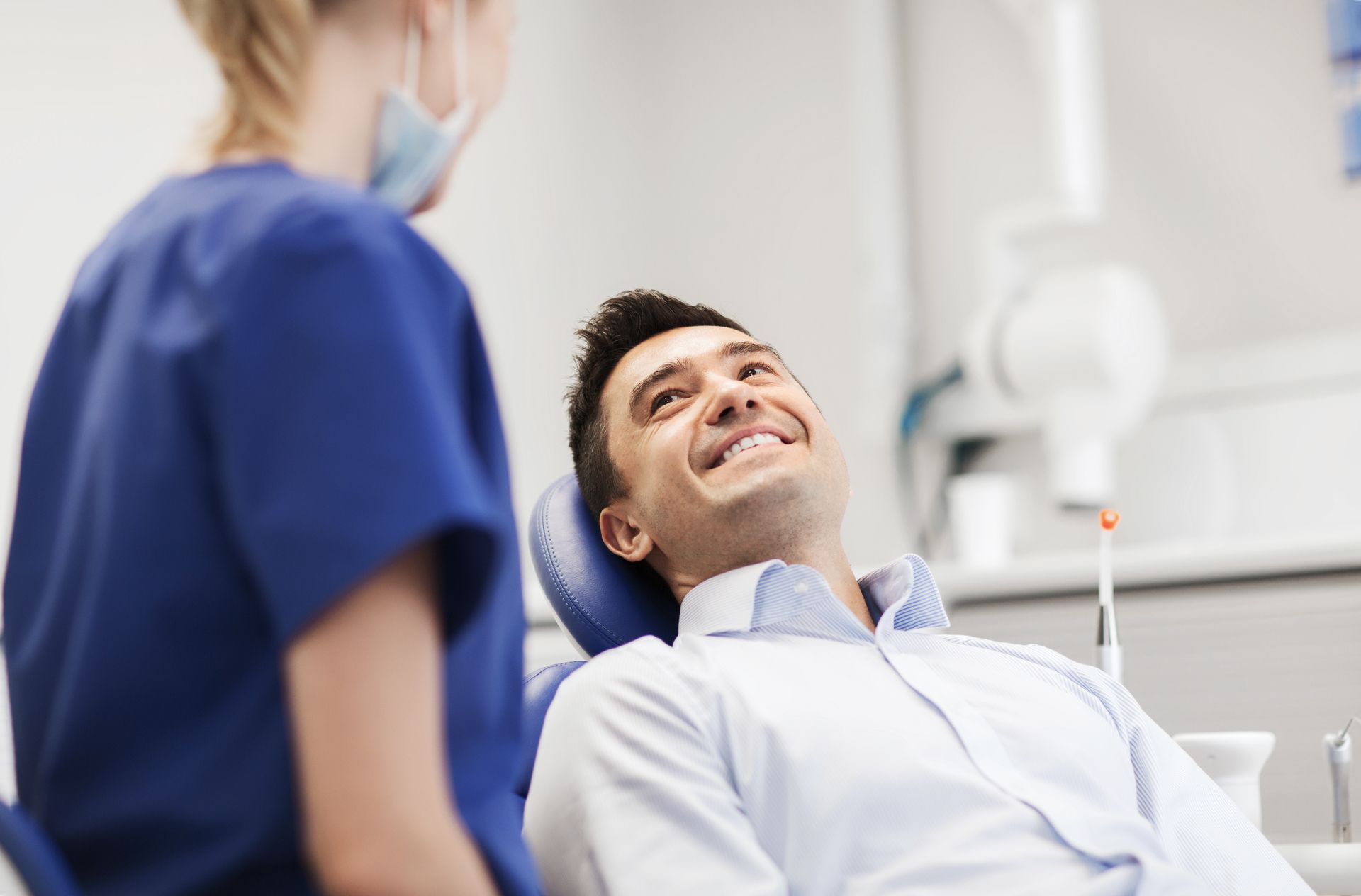 A smiling man sits in a dental chair looking up at a healthcare professional in a blue uniform.