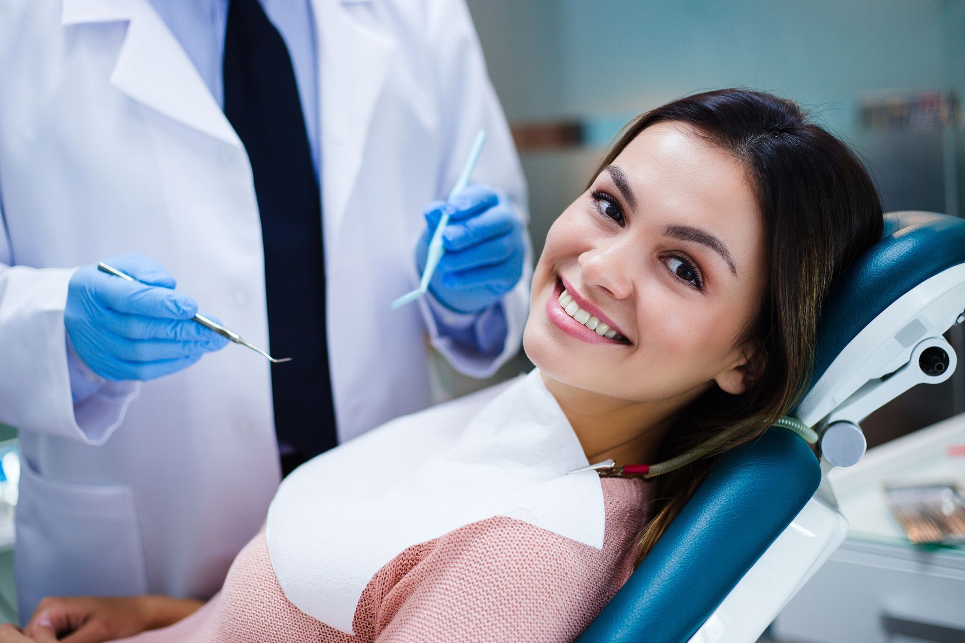 A smiling person sits in a dentist’s chair while a professional in a lab coat and blue gloves holds dental instruments.
