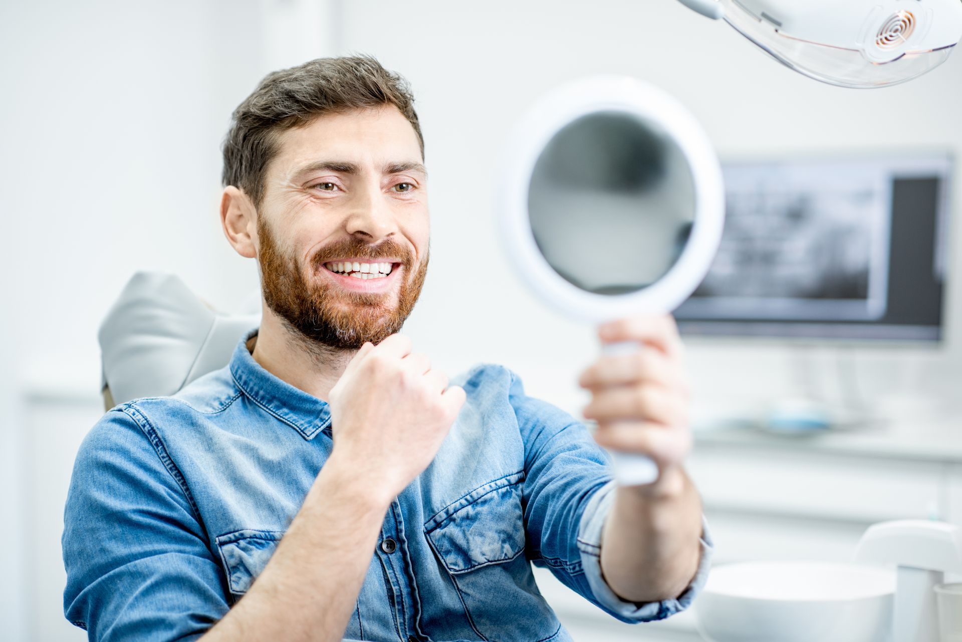 A bearded person in a denim shirt sits in a dentist's chair, smiling and looking into a hand mirror.