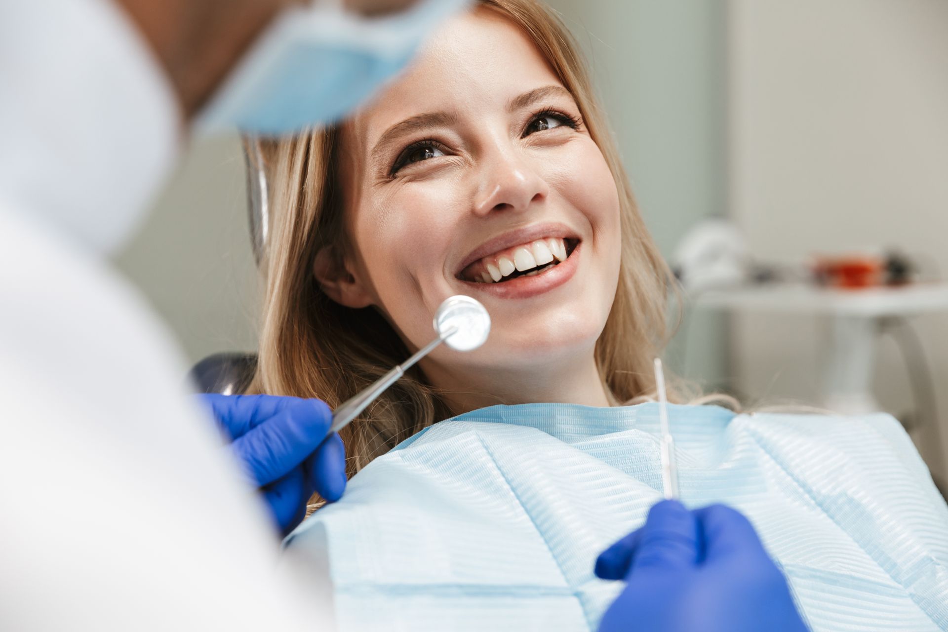 A dental professional wearing blue gloves holds a mirror to examine a smiling patient in a dental chair.
