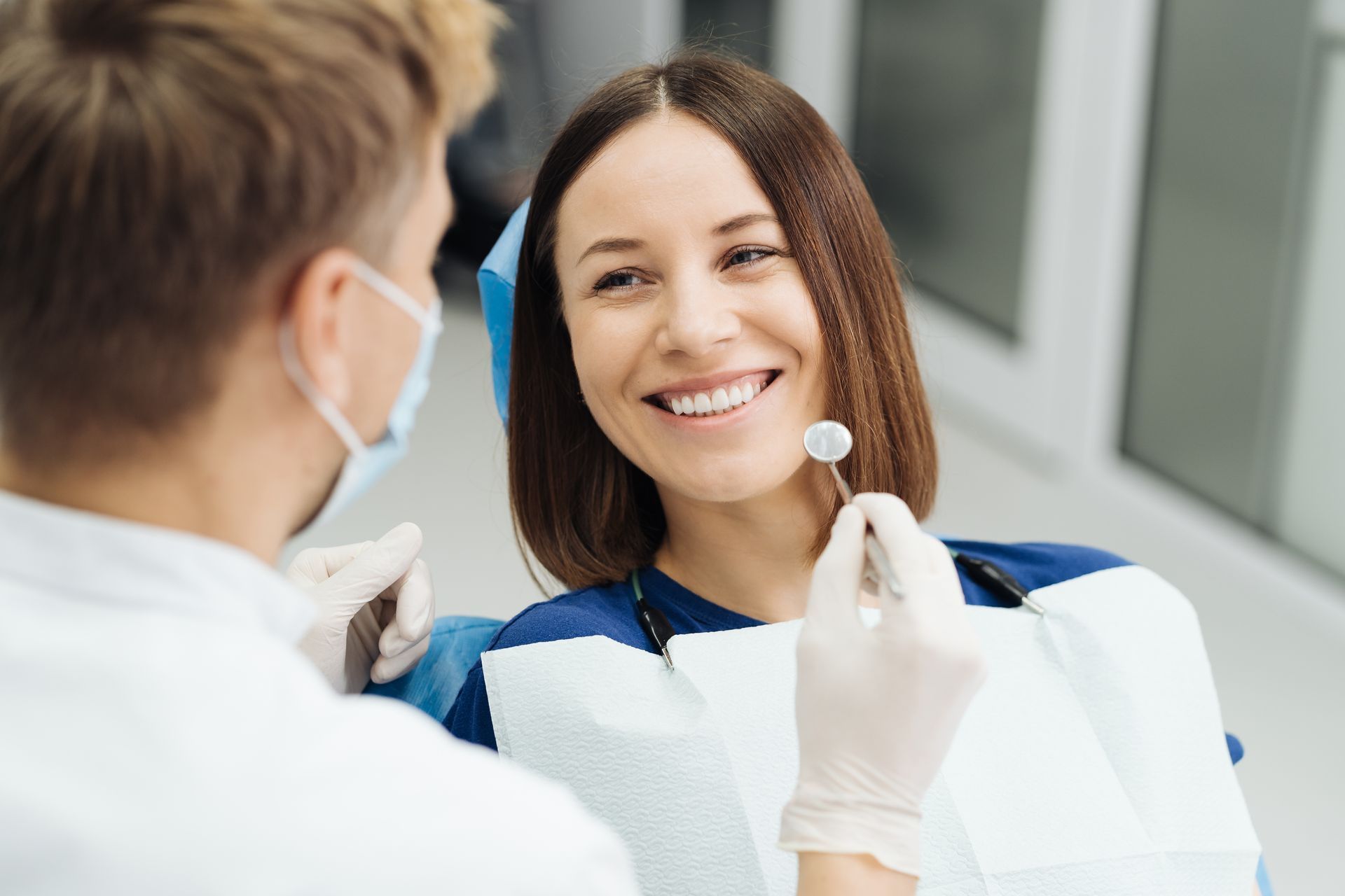 A dental professional wearing a mask and gloves holds a dental mirror for a patient smiling in a clinic setting.