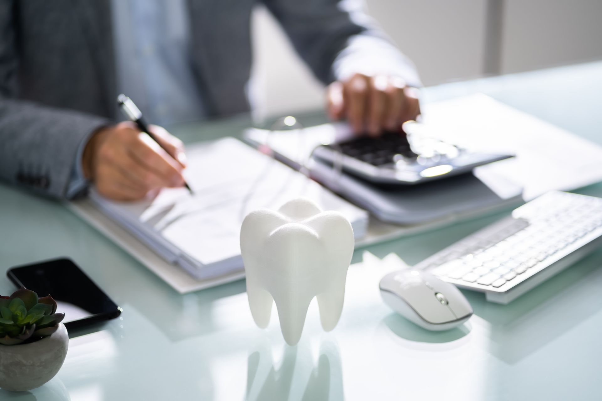 A person in a suit works at a desk with a white 3D tooth model, a calculator, a notebook, and a computer mouse.