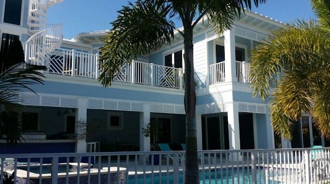 Two-story blue and white house with balcony overlooking a pool, palm trees in front.