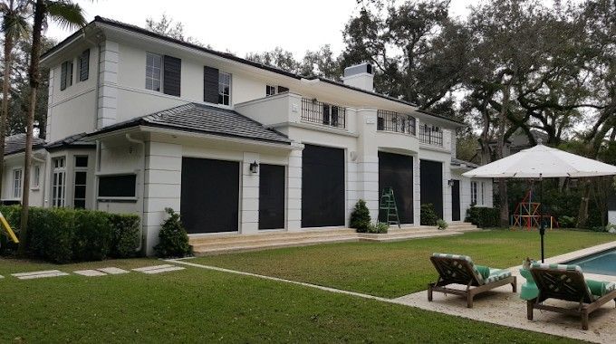 Two-story white house with black shutters, pool, green lawn, and two lounge chairs.