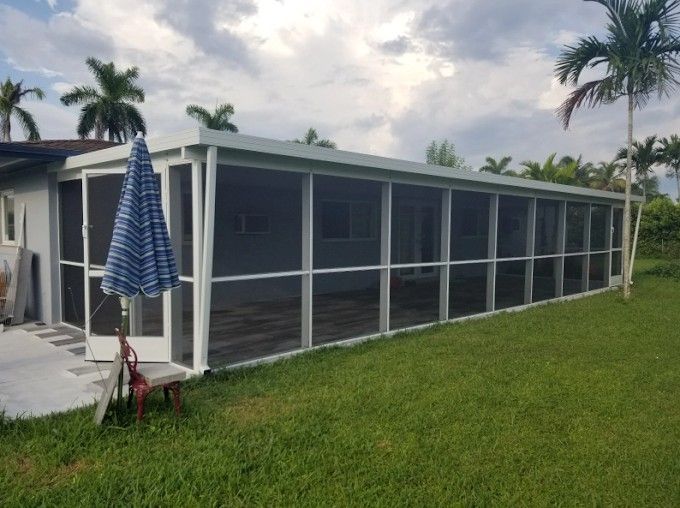 Screened porch on green grass, with blue and white umbrella, palm trees and cloudy sky.