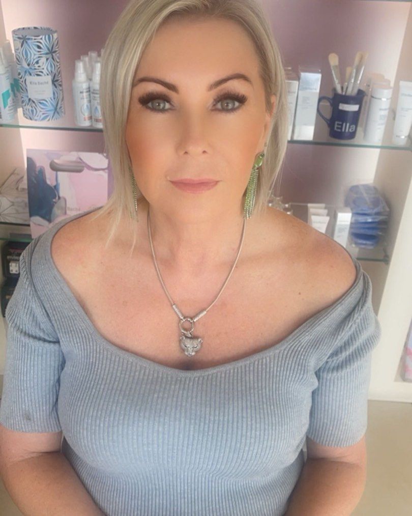 Blonde woman with makeup, wearing a gray off-shoulder shirt and a silver pendant, in a beauty setting.
