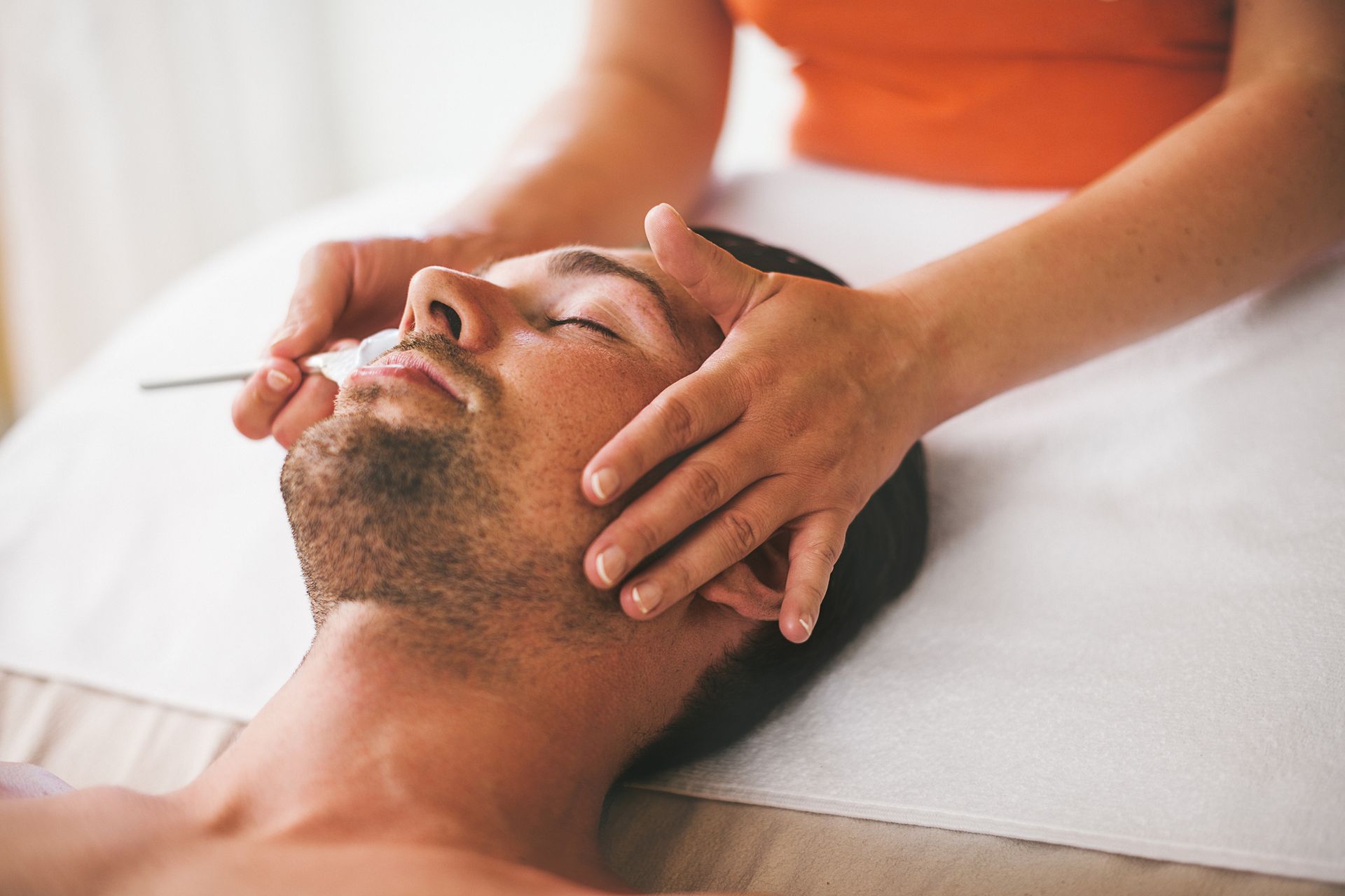 Man receiving a facial massage; masseuse's hands on his face in a well-lit room.