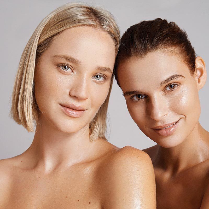 Two women with fair skin, smiling, and shoulder to shoulder against a neutral background.