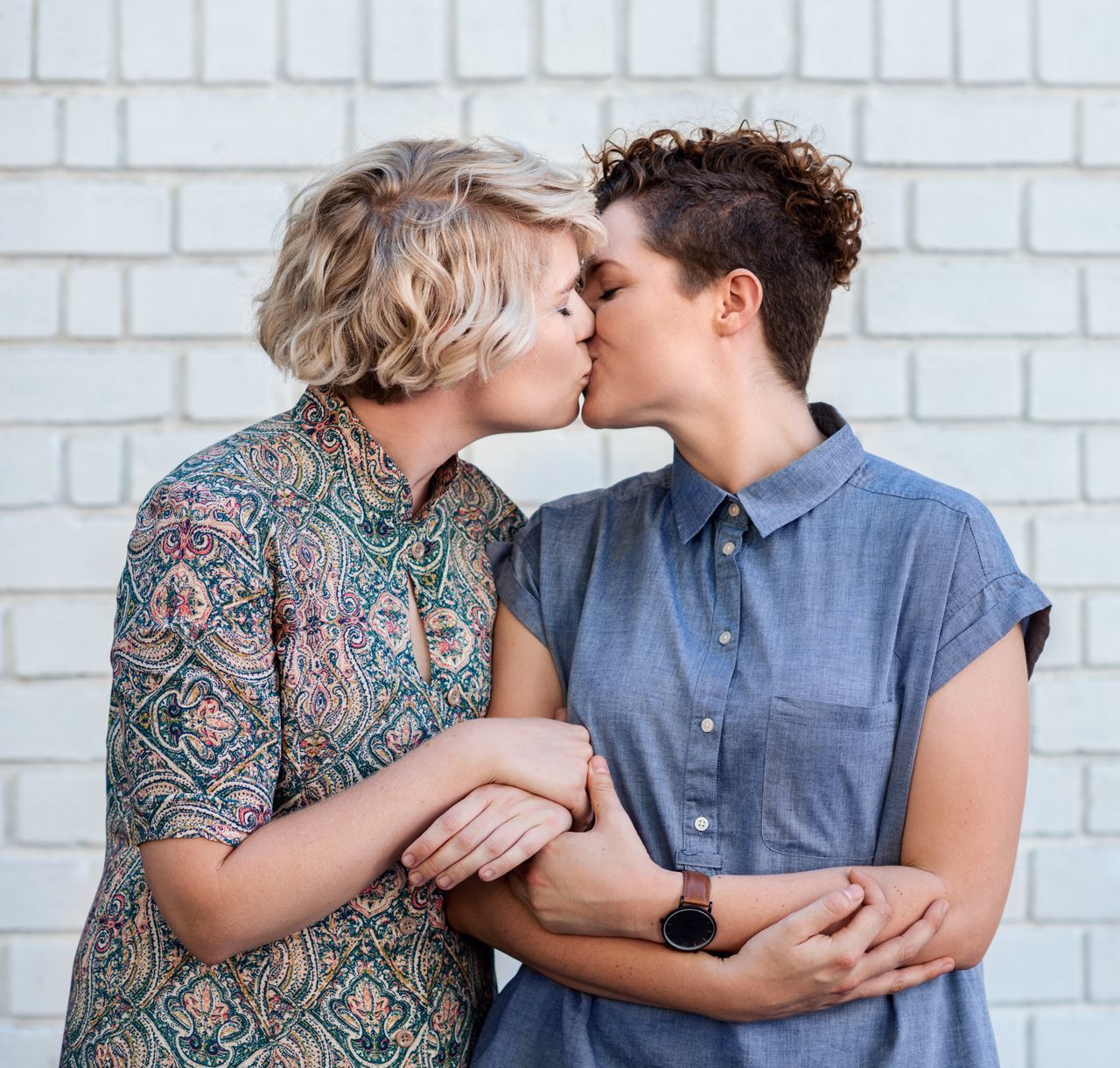 Two women are kissing in front of a white brick wall.