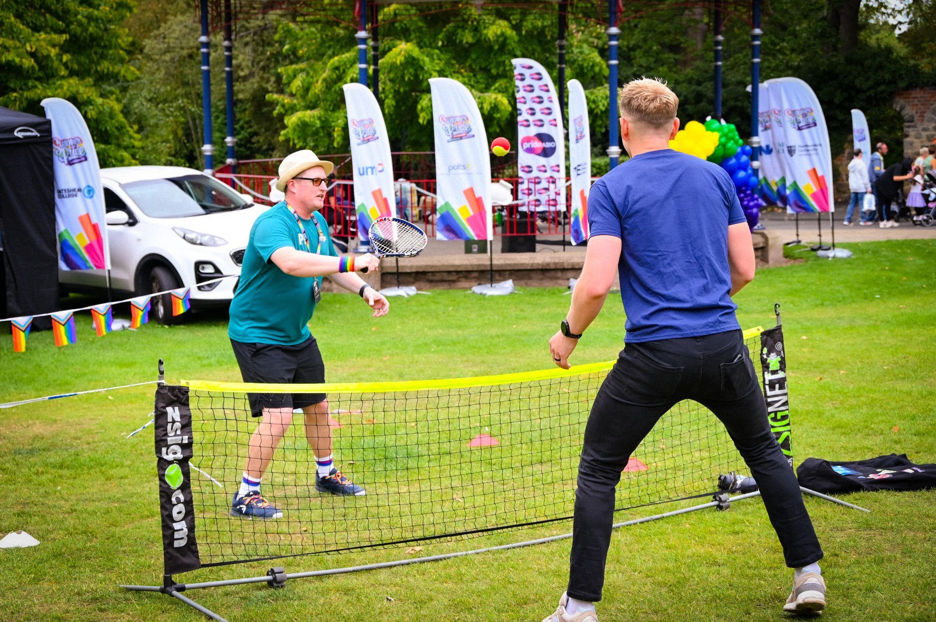 People at an outdoor Pride event cheer, some holding rainbow flags and flags with other colors, near a metal barrier.