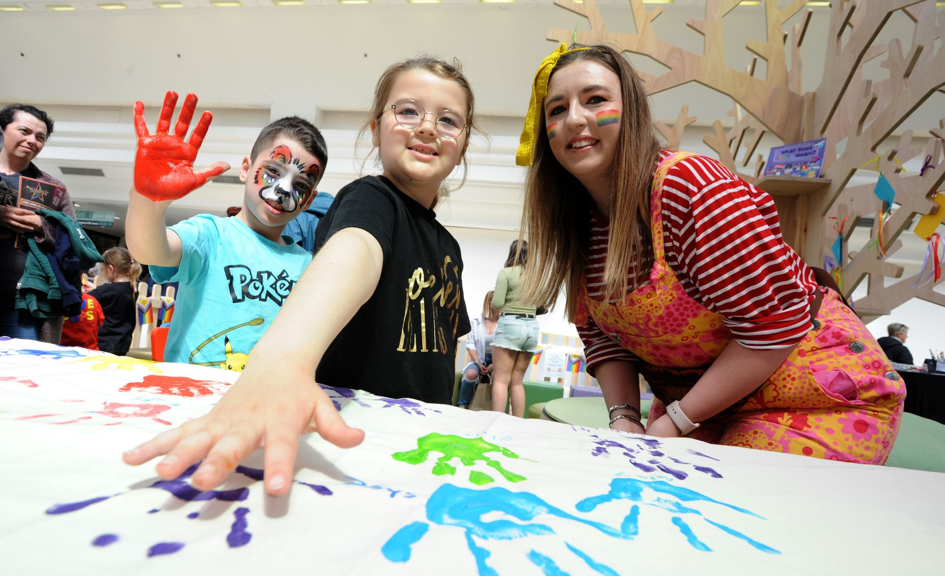 Children and an adult make handprint art at an event. The boy waves a hand with red paint.