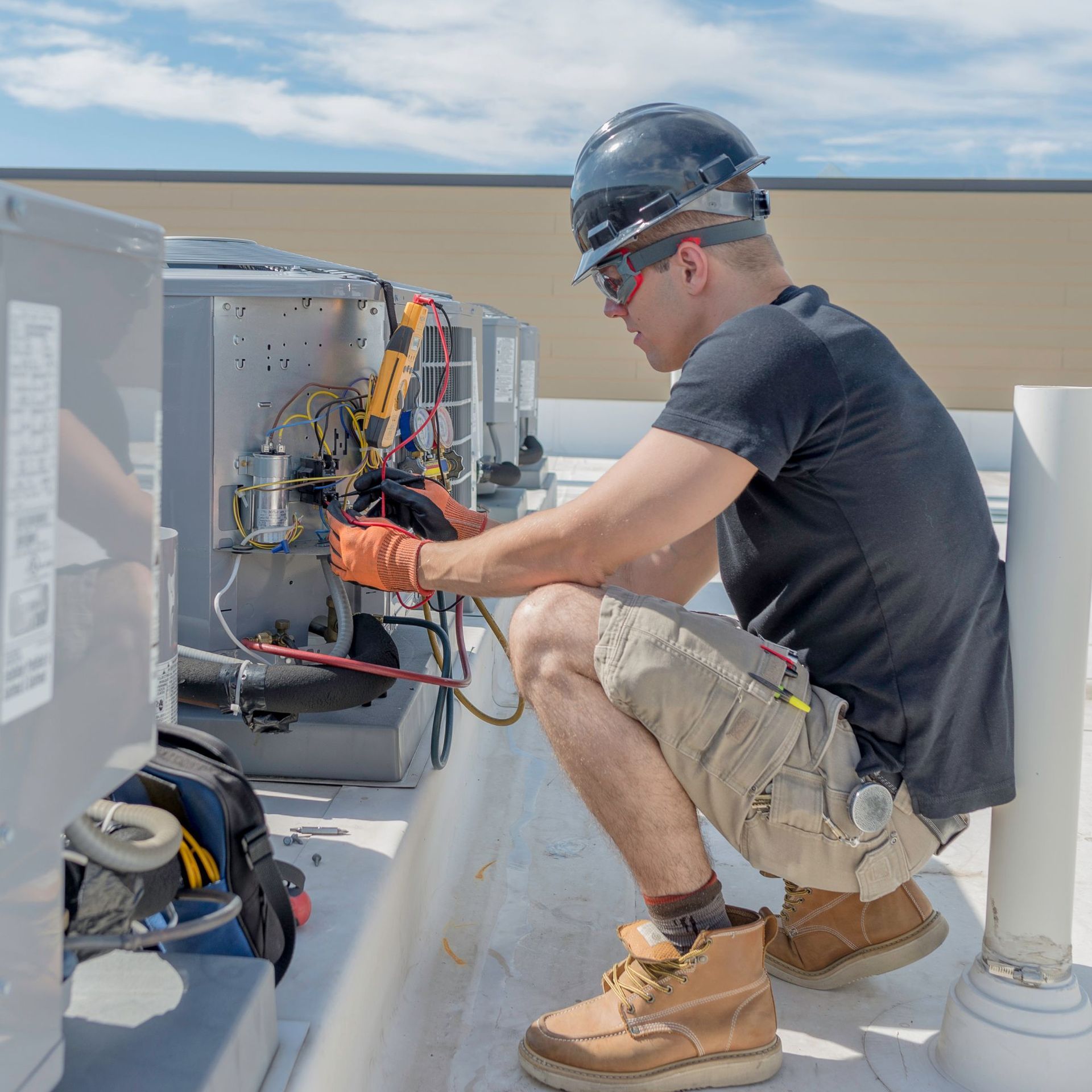 Above Comfort Mechanical LLC - HVAC technician wearing safety gear works on rooftop AC unit, testing wires with a multimeter.