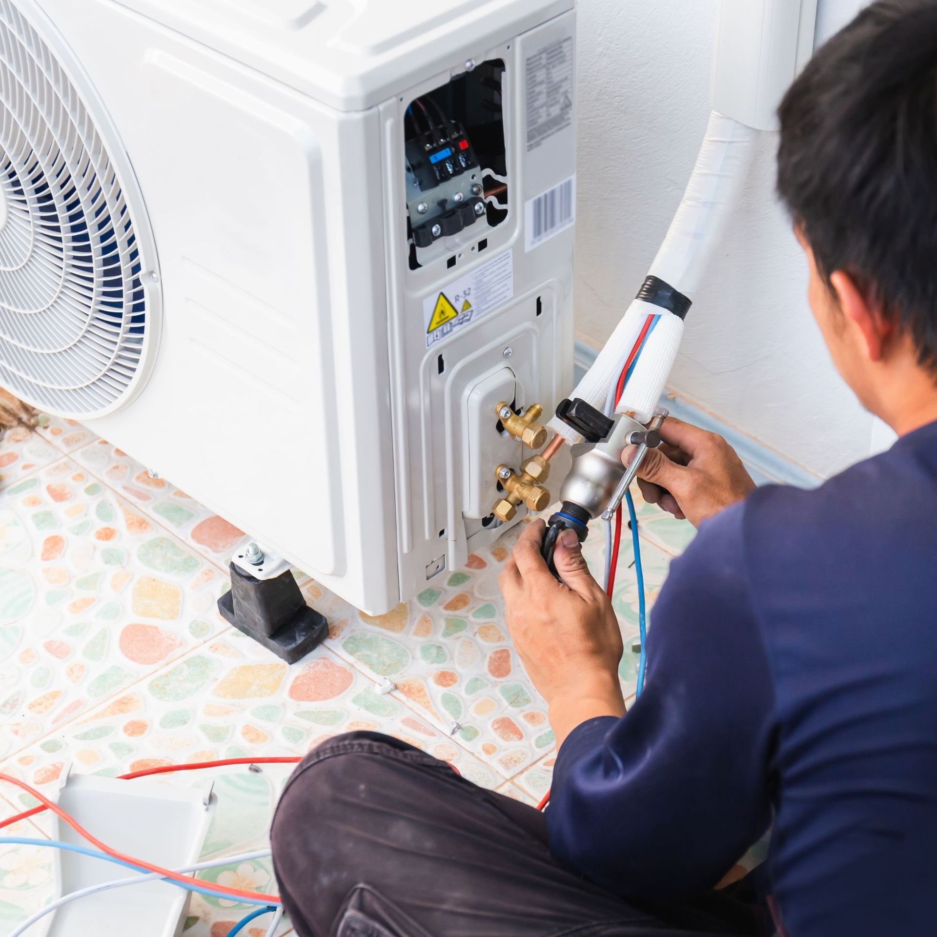 Man installing an air conditioner unit, using tools and connecting pipes outdoors.