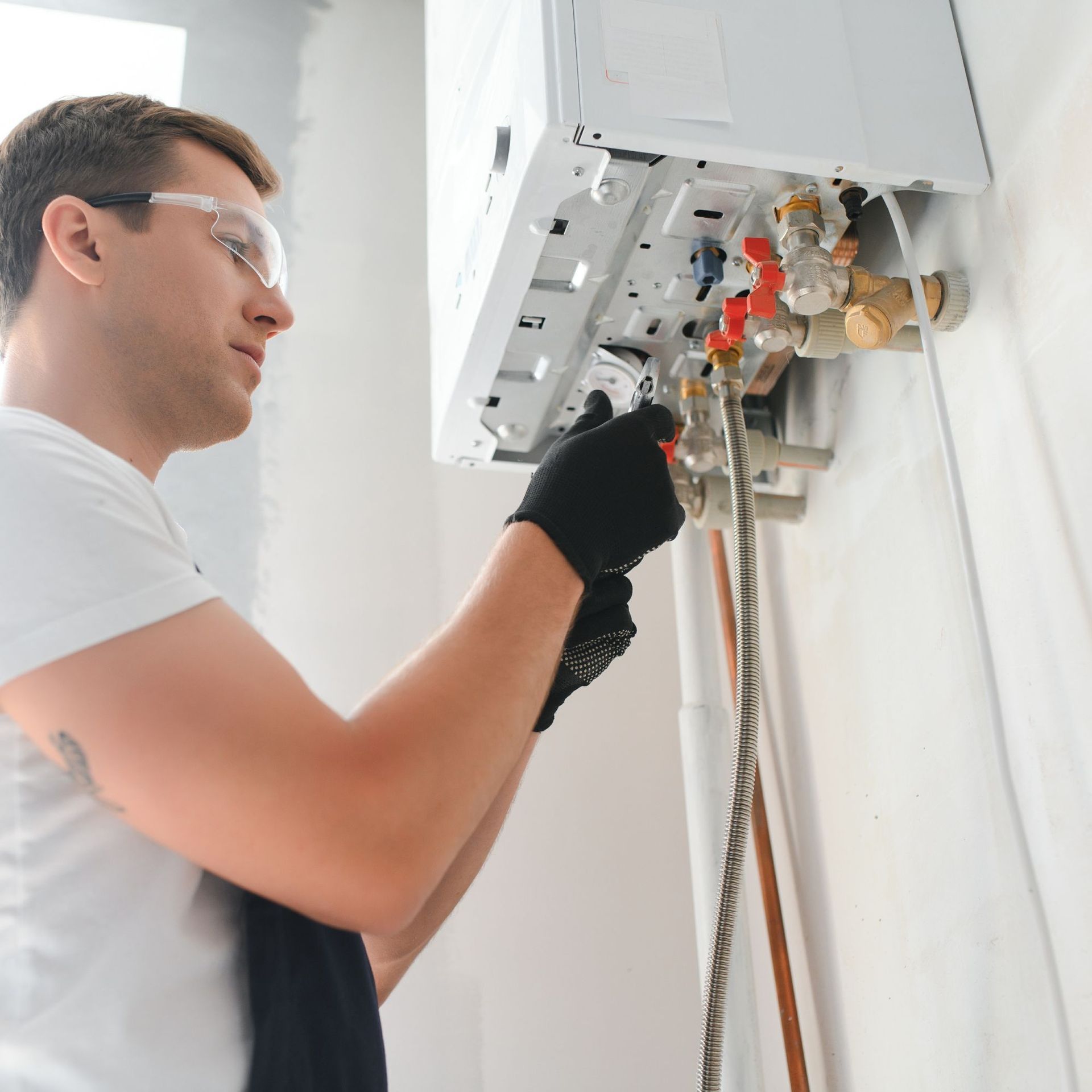 Above Comfort Mechanical LLC - Man in safety glasses and gloves repairs a boiler on a white wall.