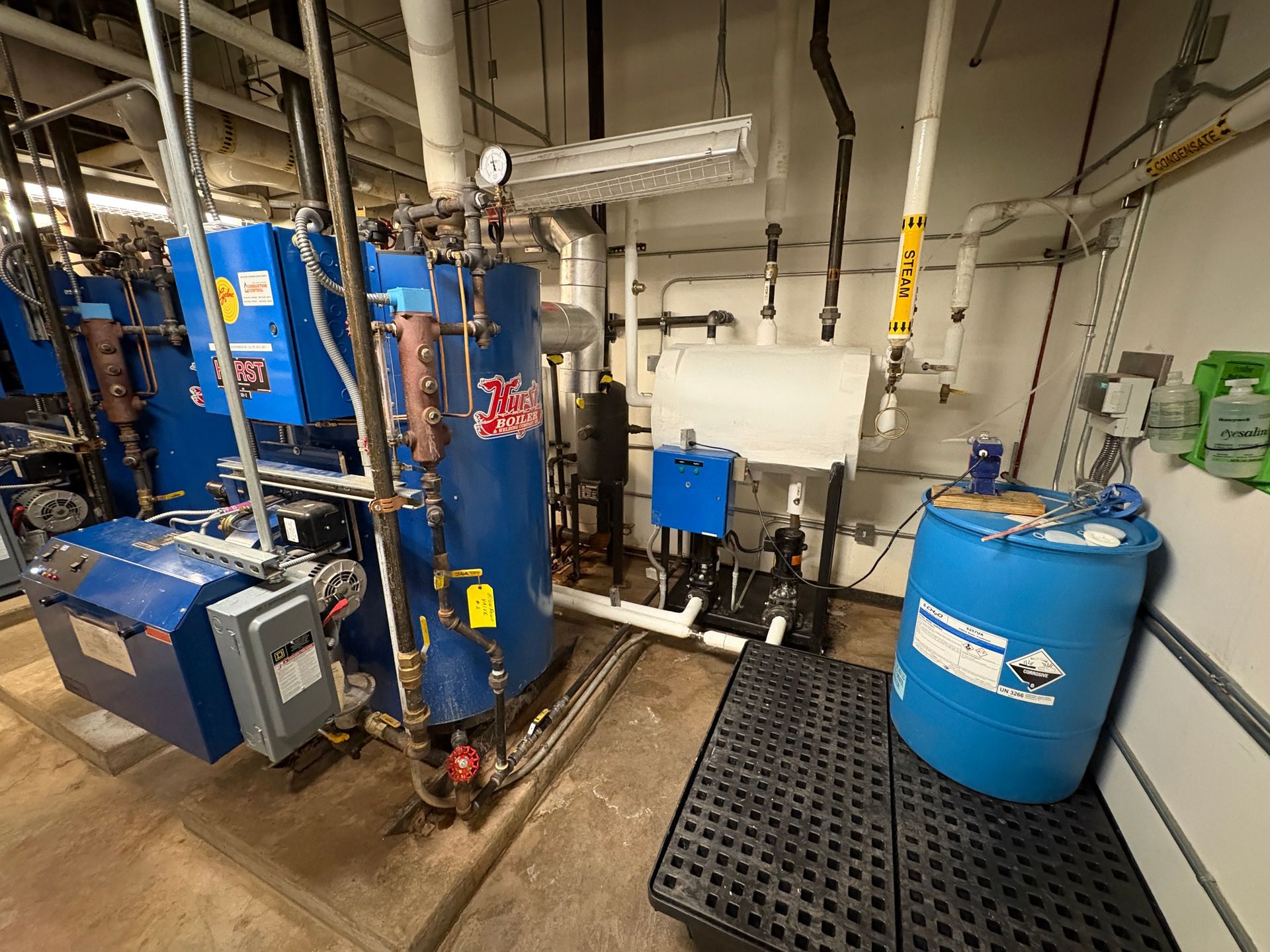 Industrial boiler room with blue machinery, pipes, and a barrel on a black grate.