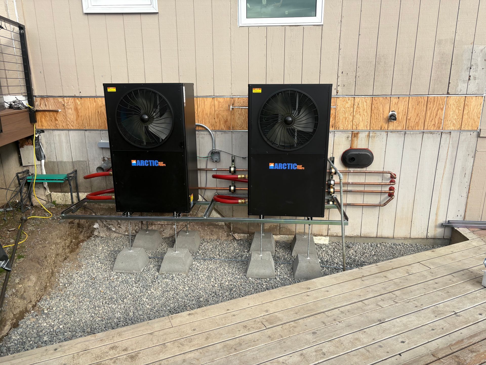 Two black air conditioning units mounted on a wall, with fan grills and red pipes, outdoors.